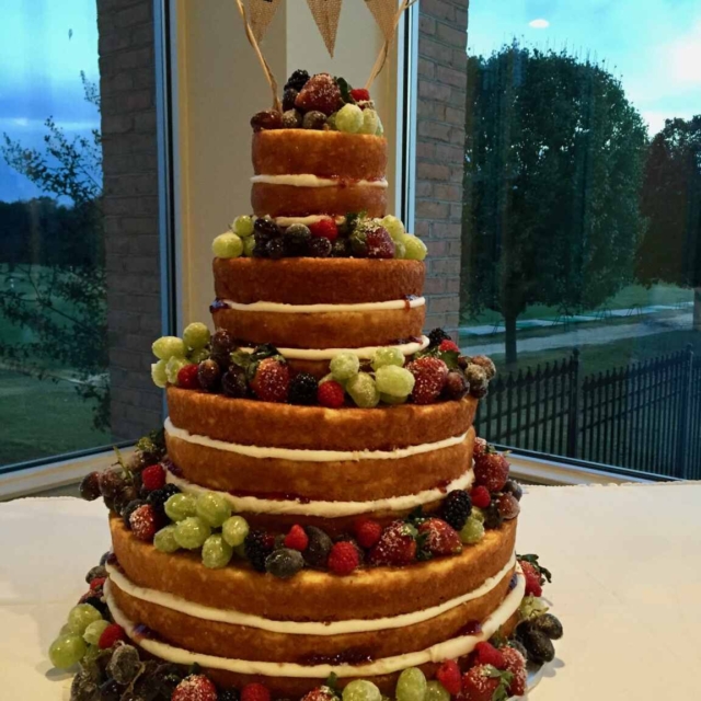 4 tier round wedding cake that has 3 layers of cake for the bottom 2 tiers and 2 layers of cake for the top 2 ties. Layers are separated by white icing, while the sponge cake is left visible. Each tier is surrounded by a selection of fruits including, Strawberries, Grapes, Blueberries, and Raspberries.