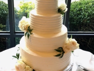 4-tier-white-wedding-cake-horiontal-groove-icing-white-roses 4 tier round wedding cake iced with cream colored frosting in a tracked pattern. The cake is decorated with white roses.