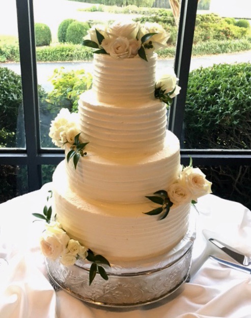 4 tier round wedding cake iced with cream colored frosting in a tracked pattern. The cake is decorated with white roses.