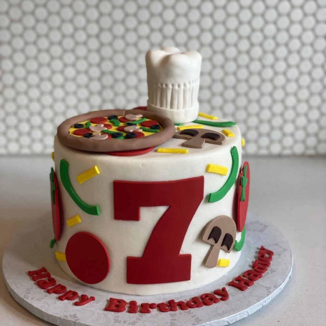 Round white cake in a chef or pizzeria design. The top of the cake includes a pizza made entirely from fondant with toppings of mushrooms, olives, bell peppers, and pepperoni. The top also has a 3d Chef's hat that is also made from fondant. The sides of the cake are decorated with fondant pizza toppings, as if they've been sprinkled down the sides, and also has a fondant number 7. The message Happy Birthday Hadley is spelled out using fondant on the cake board.