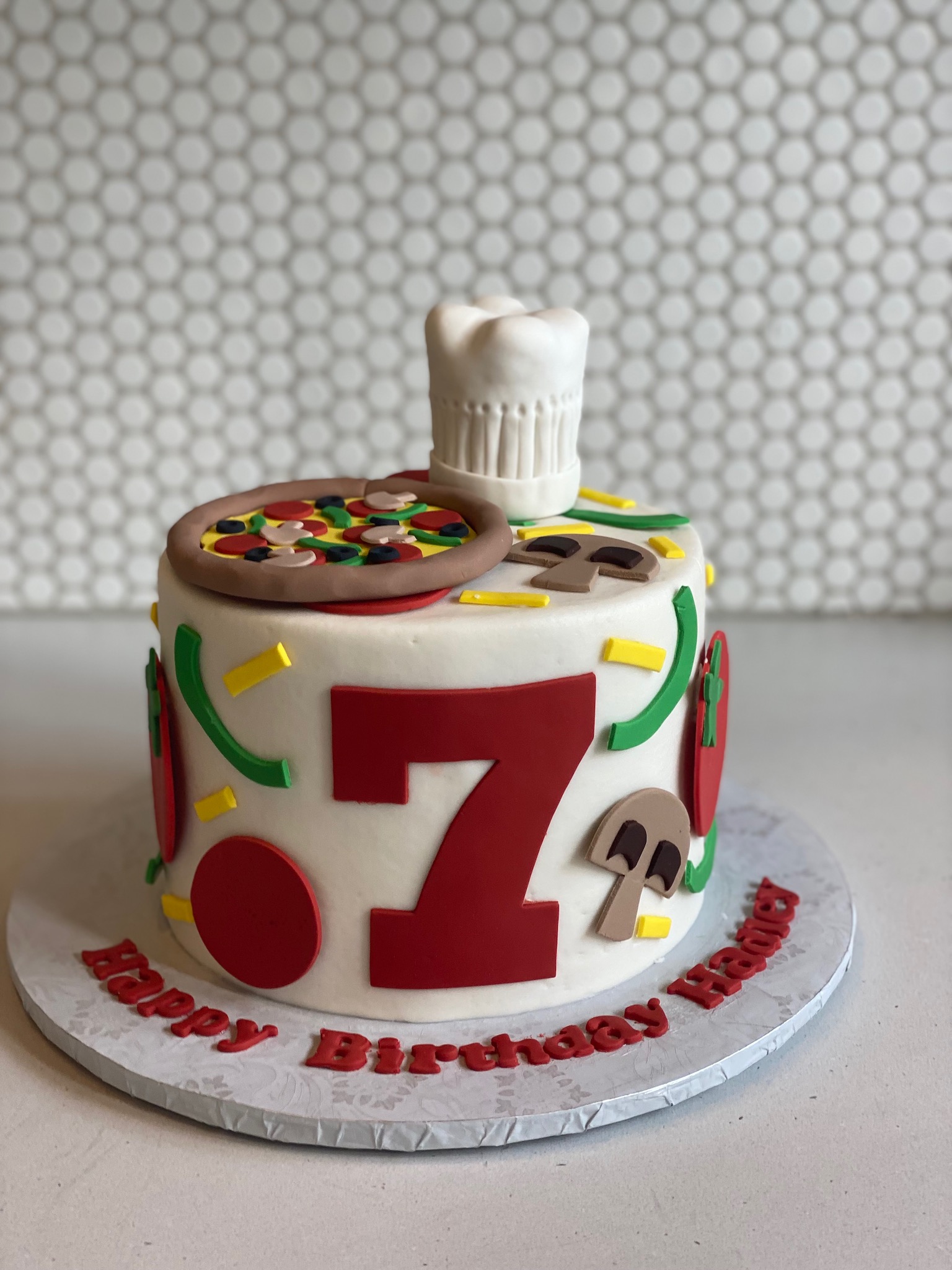 Round white cake in a chef or pizzeria design. The top of the cake includes a pizza made entirely from fondant with toppings of mushrooms, olives, bell peppers, and pepperoni. The top also has a 3d Chef's hat that is also made from fondant. The sides of the cake are decorated with fondant pizza toppings, as if they've been sprinkled down the sides, and also has a fondant number 7. The message Happy Birthday Hadley is spelled out using fondant on the cake board.