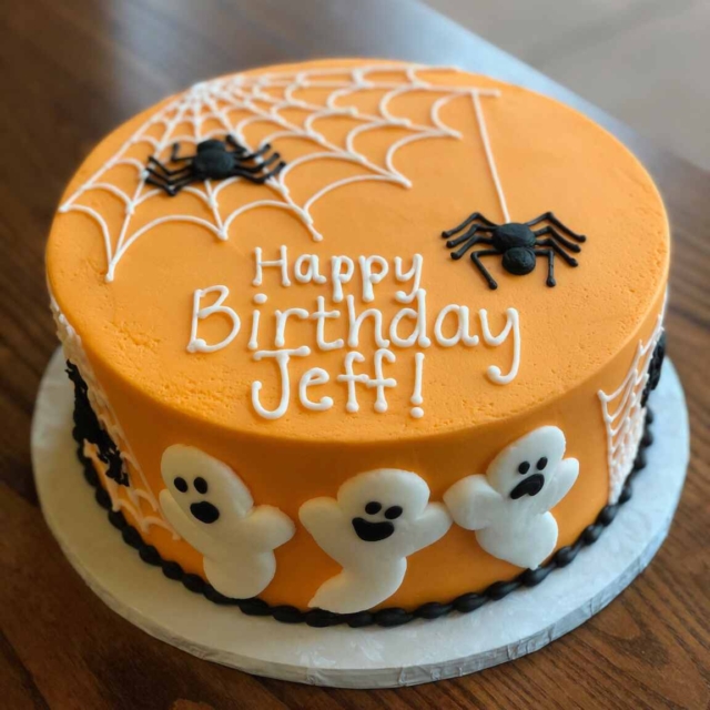 Round pumpkin colored birthday cake decorated with white fondant ghosts and black and white piped spiders with webs. The bottom of the cake is edged with piped black icing and the top of the cake has the message, Happy Birthday Jeff! in piped white icing.