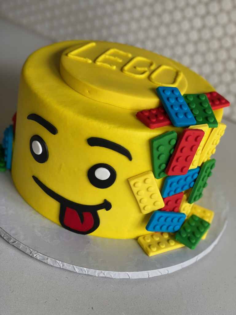 Round yellow lego cakes with lego blocks cascading down the side. Cake has a smiley face with tongue sticking out, looking like it's ready to eat a delicious slice of cake.