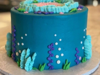 Ocean blue round birthday cake decorated with seabed vegetation in green, light blue and purple colors. The design shows bubbles rising towards the surface in white piped icing.
