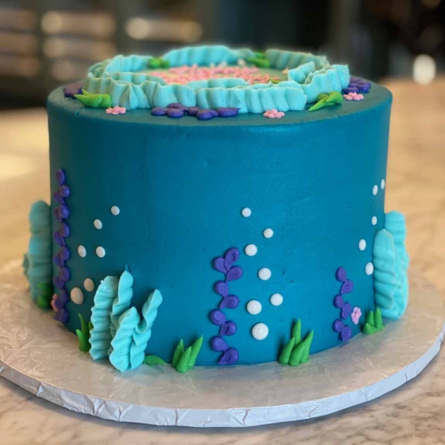 Ocean blue round birthday cake decorated with seabed vegetation in green, light blue and purple colors. The design shows bubbles rising towards the surface in white piped icing.