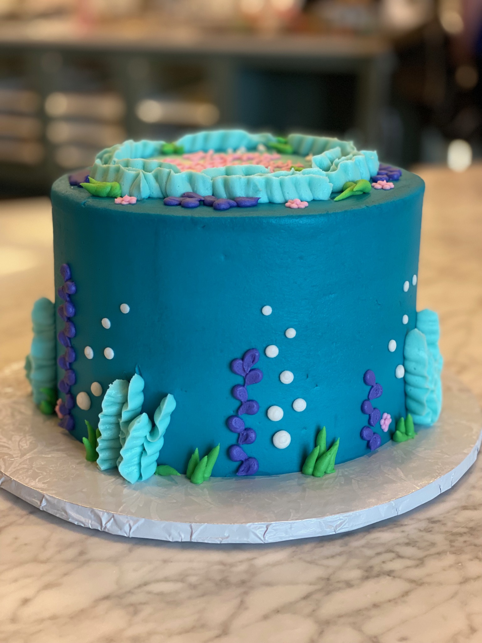 Ocean blue round birthday cake decorated with seabed vegetation in green, light blue and purple colors. The design shows bubbles rising towards the surface in white piped icing.