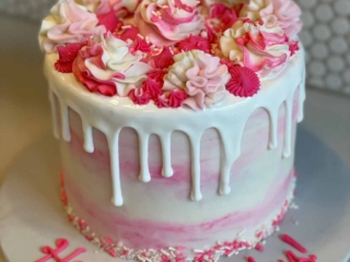 Round birthday cake intricately decorated with hand piped flowers filling the entire space of the top of the cake. Deep pinks to lighter pinks and white rose-style flower designs. The cake has the thick white icing from the top layer spilling over the edges to form a beautiful drop effect over the white and pink marbled, smoothly iced, sides of the cake. The bottom edge is finished with white and pinks sprinkles and the cake board features the piped message of Happy Birthday!