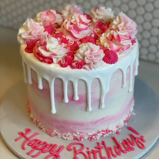 Round birthday cake intricately decorated with hand piped flowers filling the entire space of the top of the cake. Deep pinks to lighter pinks and white rose-style flower designs. The cake has the thick white icing from the top layer spilling over the edges to form a beautiful drop effect over the white and pink marbled, smoothly iced, sides of the cake. The bottom edge is finished with white and pinks sprinkles and the cake board features the piped message of Happy Birthday!