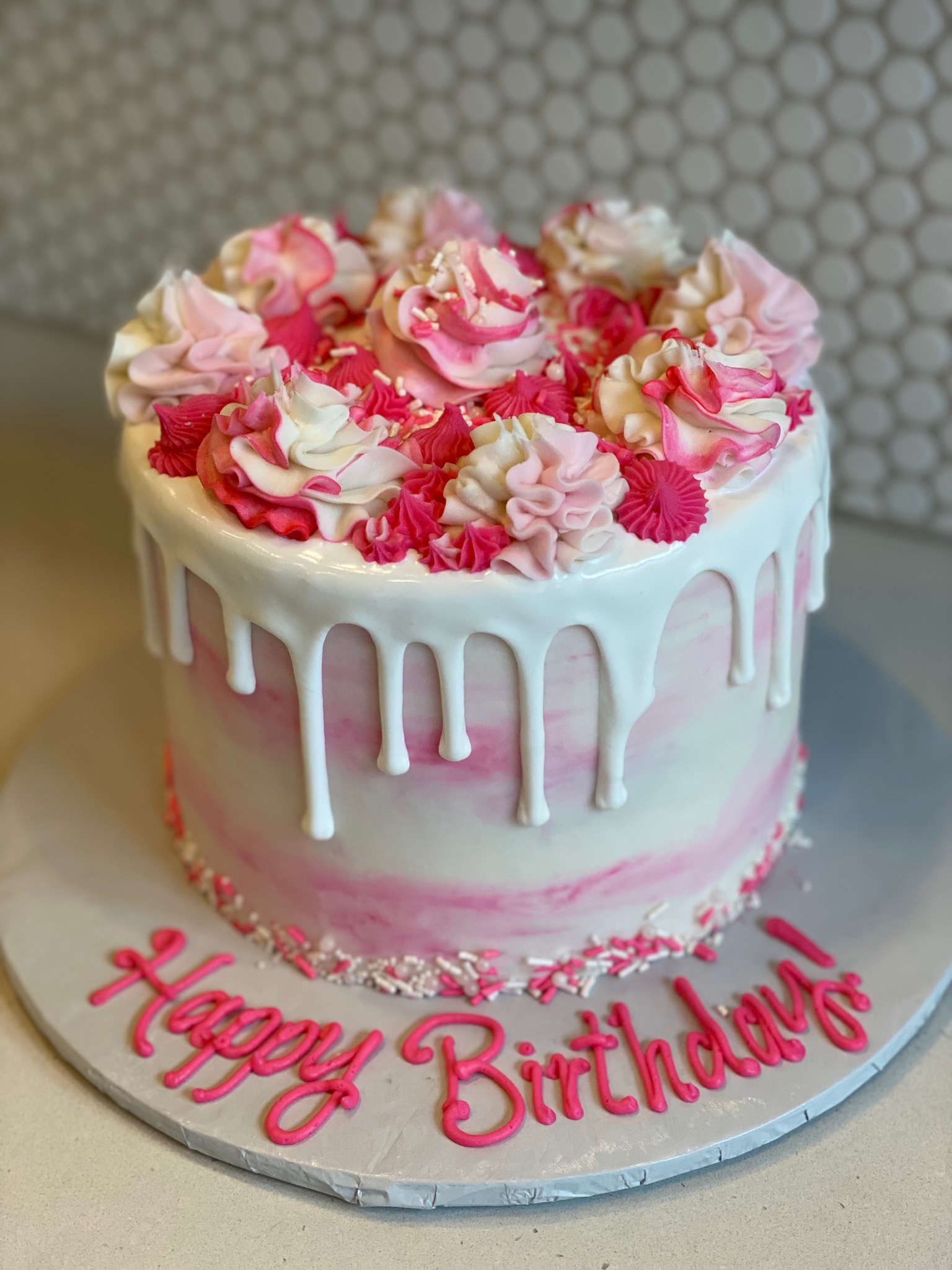 Round birthday cake intricately decorated with hand piped flowers filling the entire space of the top of the cake. Deep pinks to lighter pinks and white rose-style flower designs. The cake has the thick white icing from the top layer spilling over the edges to form a beautiful drop effect over the white and pink marbled, smoothly iced, sides of the cake. The bottom edge is finished with white and pinks sprinkles and the cake board features the piped message of Happy Birthday!