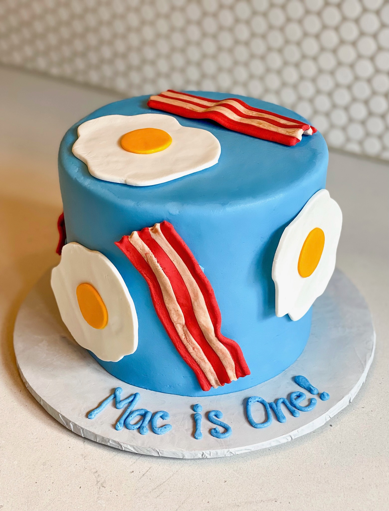 Round button blue cake with bacon and fried eggs decorations made from fondant. Cake board has piped icing with the message, Mac is one!