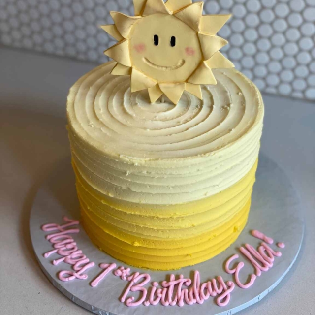 Round sun smiley birthday cake iced with graded color flowing from a vivid sunshine yellow to a pastel muted yellow. The icing is done in a grooved design and on top of the cake is a vertically standing fondant smiley faced sun, similar in design to the head of a sunflower.