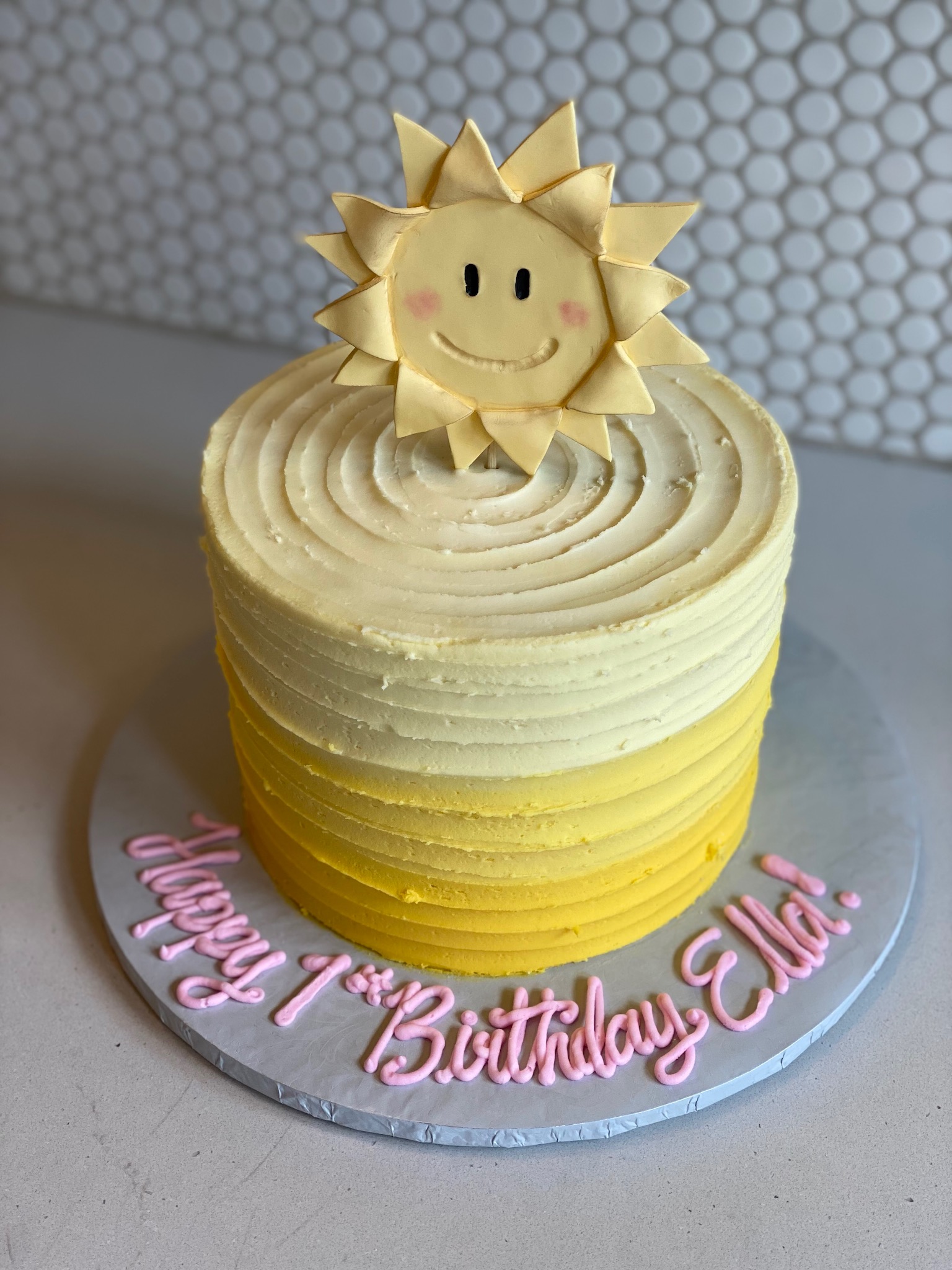 Round sun smiley birthday cake iced with graded color flowing from a vivid sunshine yellow to a pastel muted yellow. The icing is done in a grooved design and on top of the cake is a vertically standing fondant smiley faced sun, similar in design to the head of a sunflower.