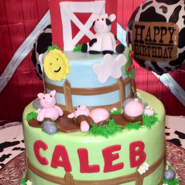 Two tier round fondant birthday cake in a farmyard design. The top layer features a classic red barn with a fondant Holstein Friesian cow sat in front of it. The top layer is blue, depicting the sky, and there is a fondant brown fence along the bottom edge. Fondant piglets are basking in a mud pool in front of the fence and on the top edge of the bottom layer. The bottom layer is green like grass along with a the name Caleb in red fondant. Finally, around the bottom edge is a duck pond, rocks, and 3 yellow fondant ducks swimming.