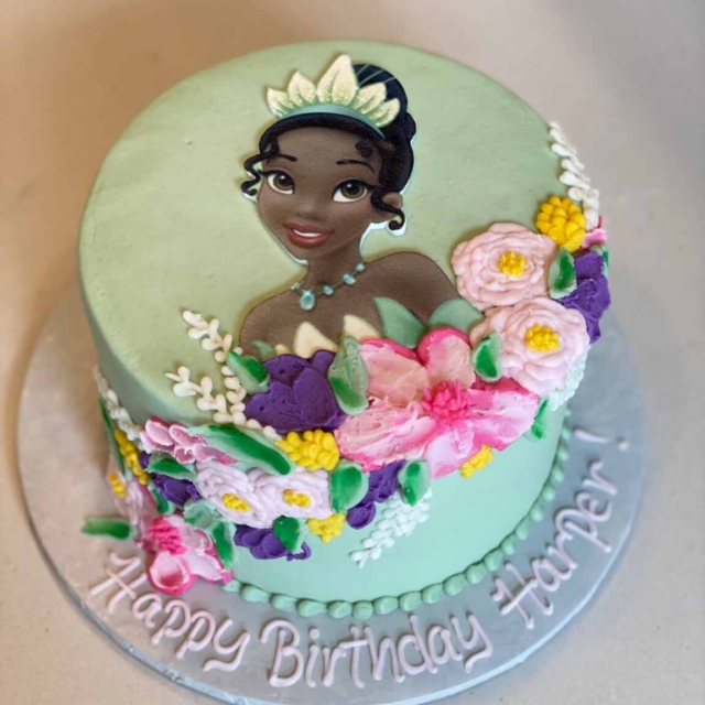 A round wintermint colored cake featuring a hand piped princess design. The cake also has piped icing in a cascading flower arrangement design. Wintermint colored piped icing pearls line the outer edge of the bottom of the cake. The cake board has a piped Happy Birthday Harper message.