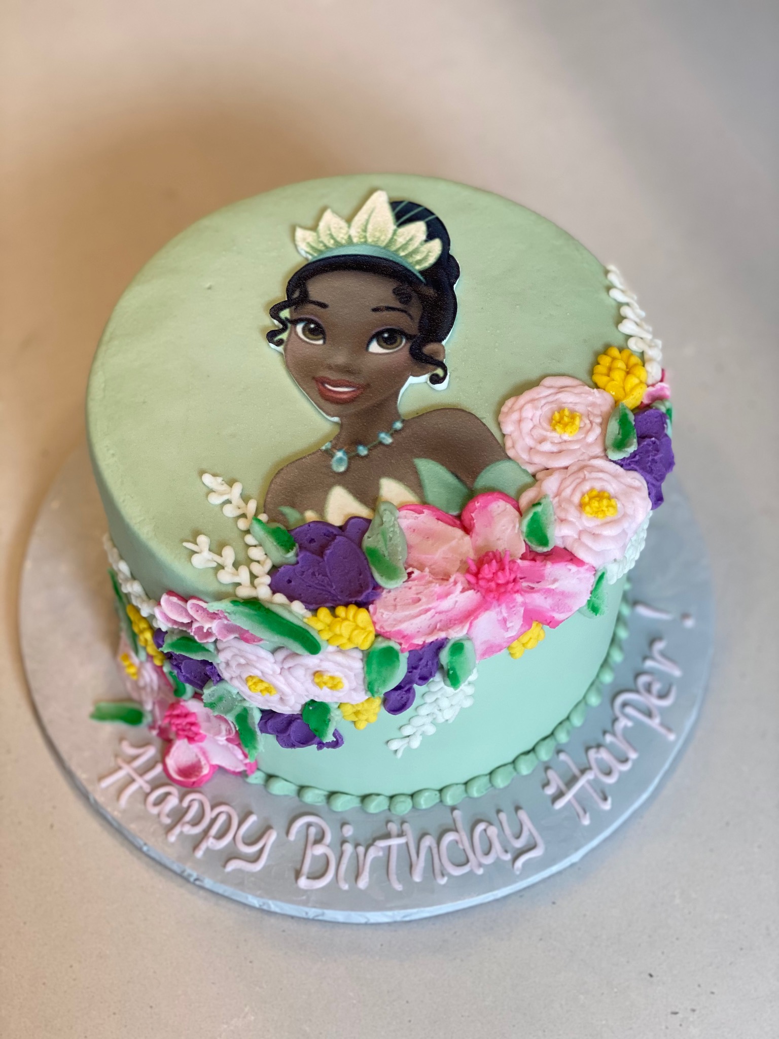 A round wintermint colored cake featuring a hand piped princess design. The cake also has piped icing in a cascading flower arrangement design. Wintermint colored piped icing pearls line the outer edge of the bottom of the cake. The cake board has a piped Happy Birthday Harper message.